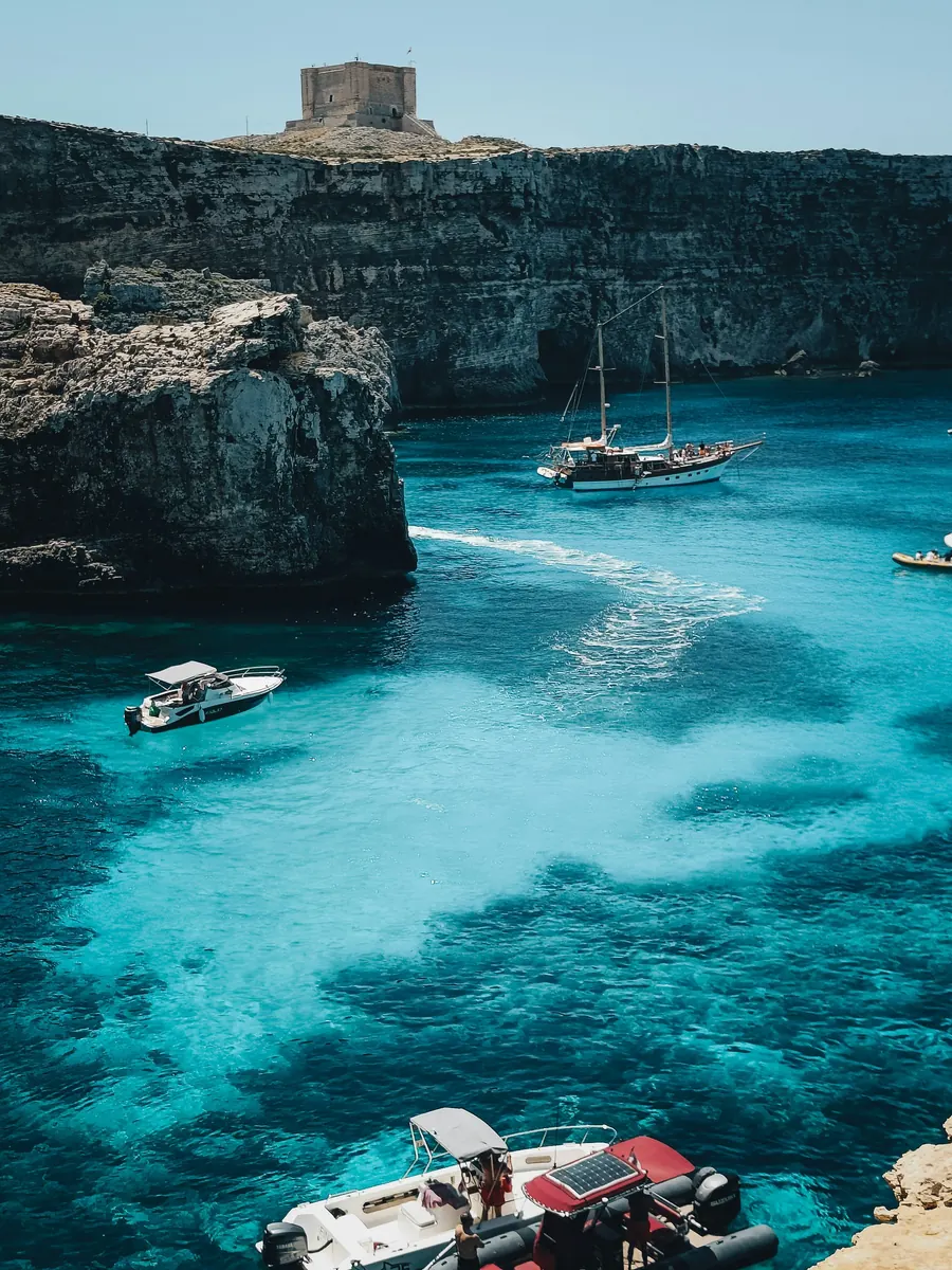 Turquoise water below limestone cliffs with a small tower on the ridge and boats in the bay
